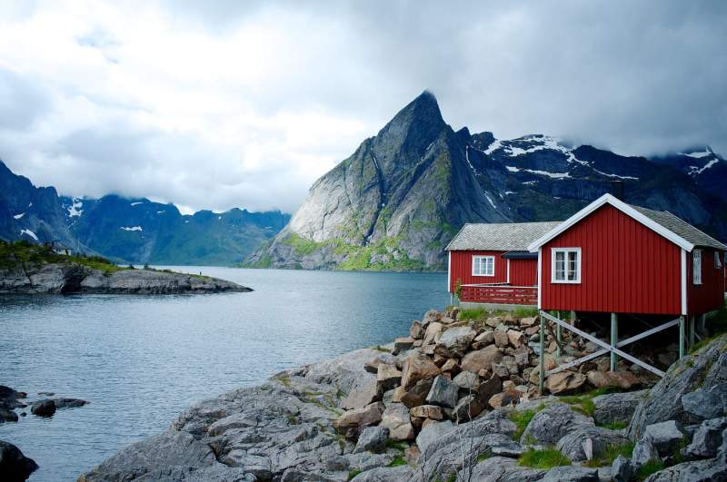 Red farmhouse built on the edge of a beautiful lake with mountains surrounding it.